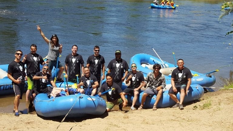 A group of people in matching tuxedo shirts sitting and standing around blue rafting boats that are tied up on the shore of a calm river. Other rafting boats full of people in the background.