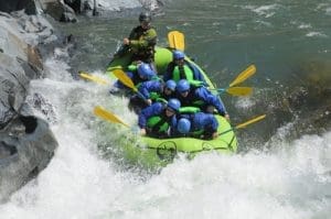 A group of people in a rafting boat paddling through a whitewater rapid. Rocks surround the river.