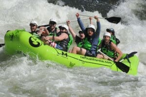 People on a green rafting boat surrounded by a whitewater rapid, holding their paddles in the air and smiling while getting splashed by the river water.