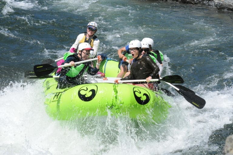 A group of people in a rafting boat paddling through a whitewater rapid. The river water is splashing the people holding paddles.