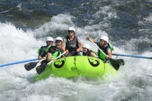 A group of people in a rafting boat paddling through a whitewater rapid. The river water is splashing the people in the boat.