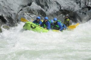 A group of people in a rafting boat navigating a large whitewater rapid. Large rock walls behind the boat.