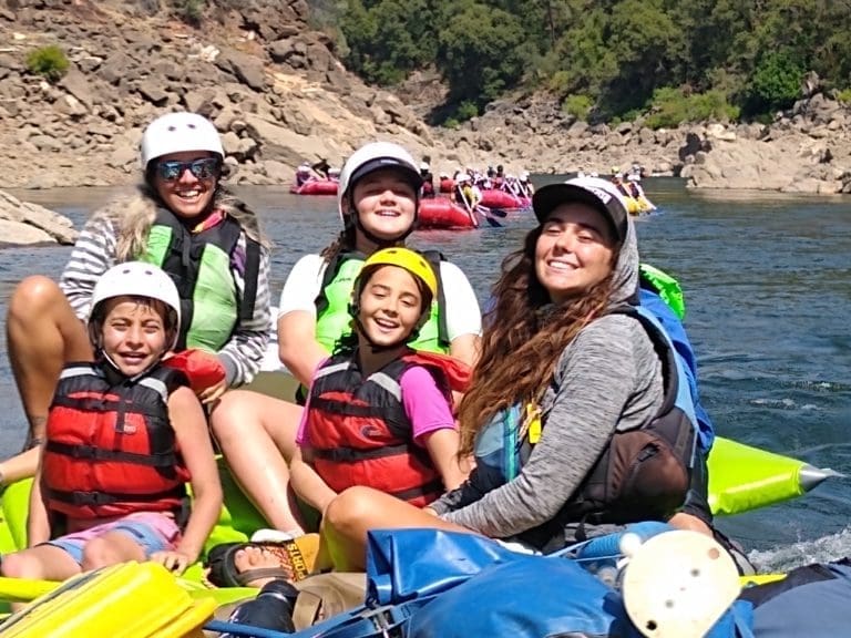 A group of people including children, sitting in a rafting boat floating down a calm river. Multiple rafting boats full of people behind them. Rocks and green trees in the background.