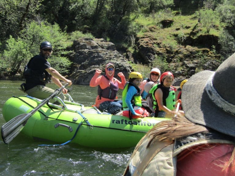 A group of people including kids on a rafting boat floating down a calm river with rocks and green trees in the background.