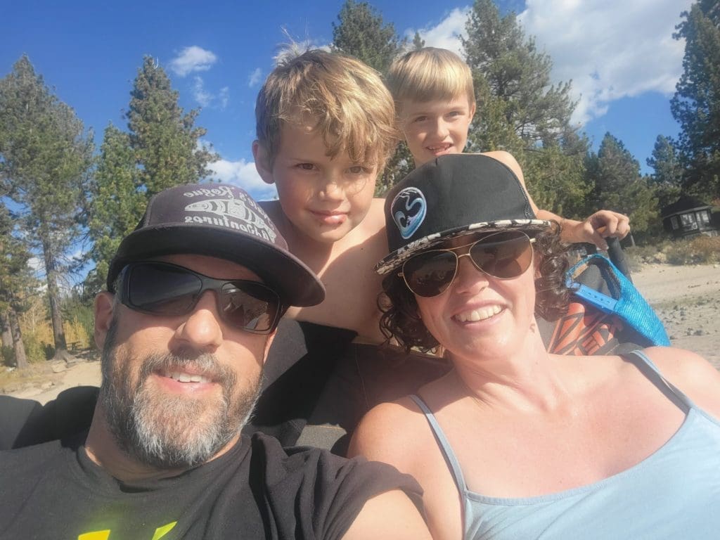 A family posing on a beach, everyone is smiling. Green trees and blue skies in the background.