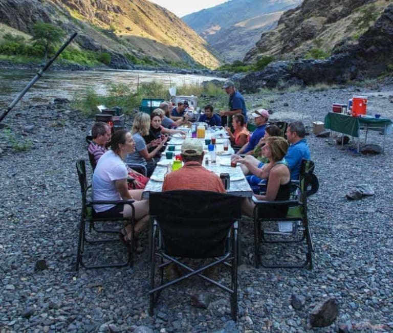 A group of people sitting around a table eating dinner at sunset. The table is on a rocky beach next to a calm river with a grassy canyon in the background.