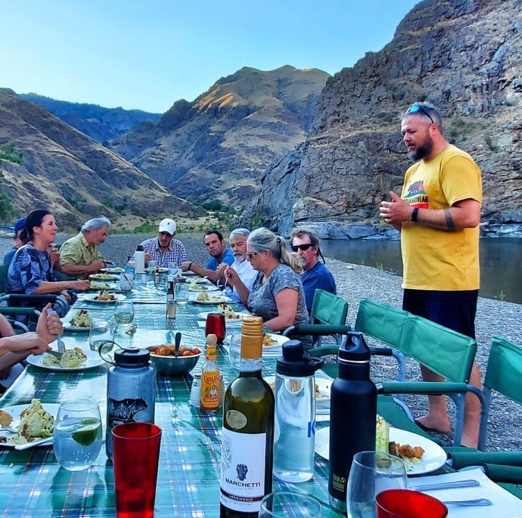 A group of people sit around a dinner table with various food and beverages on the shore of a calm river. Large rock wall canyon in the background.