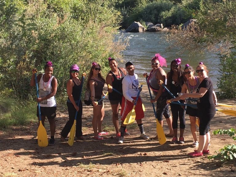 A group of people standing on the beach with a river with some whitewater behind them. They're holding paddles with matching purple hats.