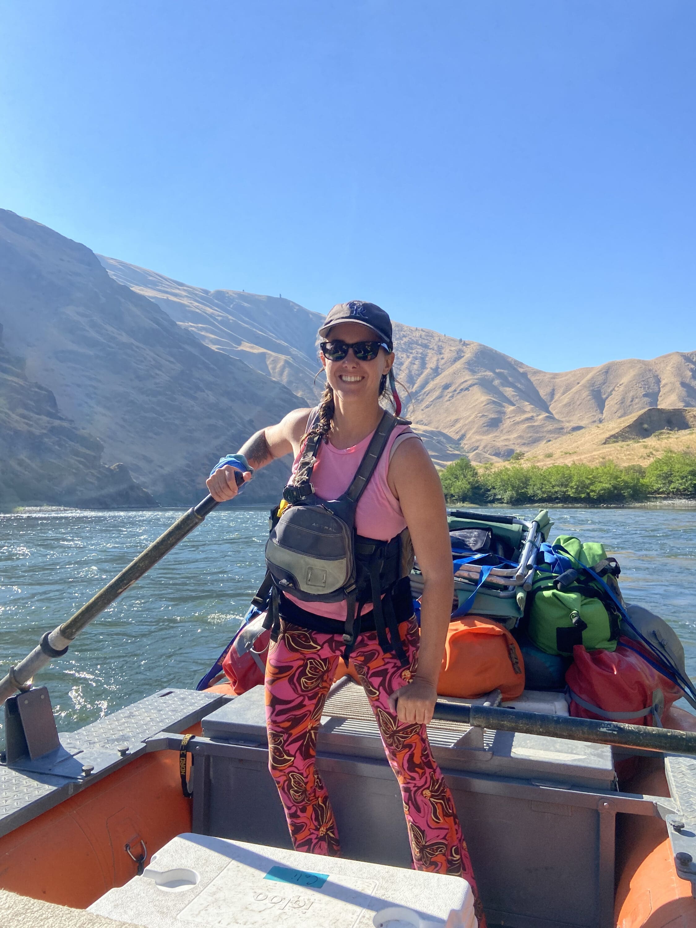 A woman standing in a rafting boat holding a paddle that is in the water. There is a pile of gear on the boat behind her. Behind her is a brown grassy hillside.