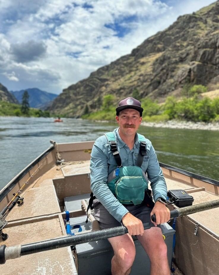 A whitewater guide sitting in a fishing boat on a calm river. Another boat is behind him floating on the river with a rocky mountain in the background.