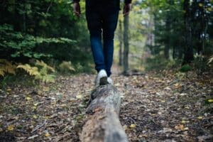 A close up picture of a long in a forest with colorful leaves on the ground and green trees in the background. A person is walking on the narrow log, showing only their legs and hands.