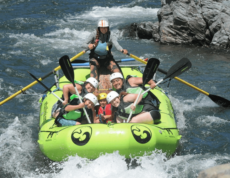 Family posing in an inflatable rafting boat with a guide sitting behind them. They're all wearing helmets and lifejackets while holding their paddles in the air. The boat is in whitewater with a large rock behind them.