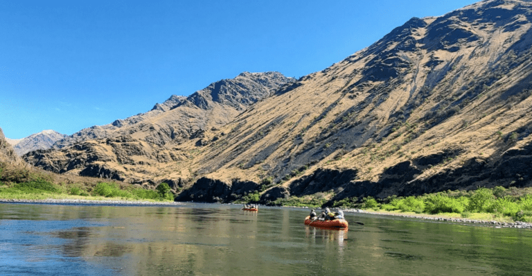 Two inflatable rafting boats floating down a calm river. The banks have bright green bushes with a tall brown hillside above it.