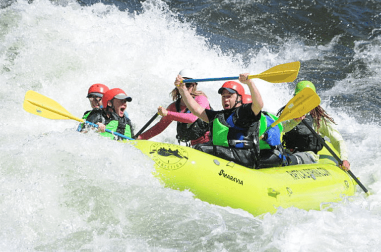 Group of people in a green inflatable rafting boat all wearing helmets and lifejackets and holding paddles. They are in the middle of a large whitewater rapid, you can't see any calm river in the photo.