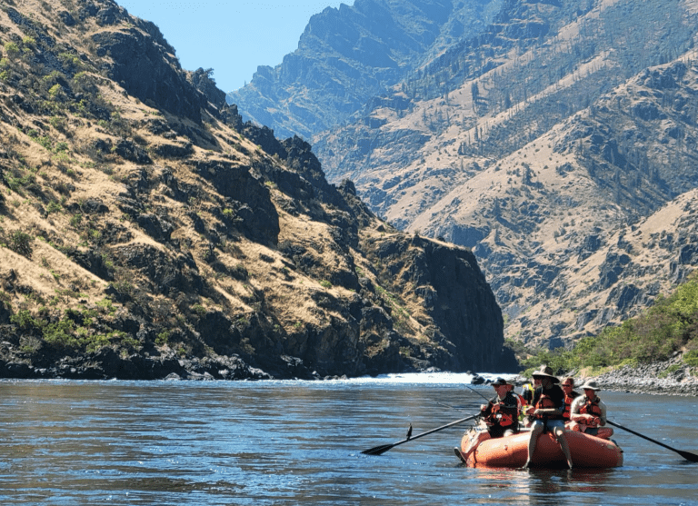 One orange inflatable rafting boat on a calm river. There is a small whitewater rapid behind them. One person is sitting on the front of the boat and another one is holding a fishing pole with a fish on the line. Large canyon walls in the background full of rocks, shrubs and trees.