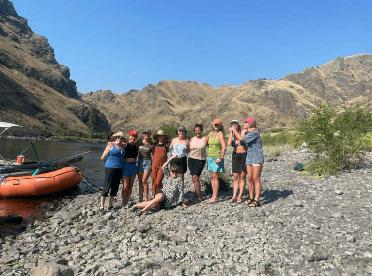 A group of women standing on the rocky shore of a river next to an orange inflatable rafting boat that is tied to shore.