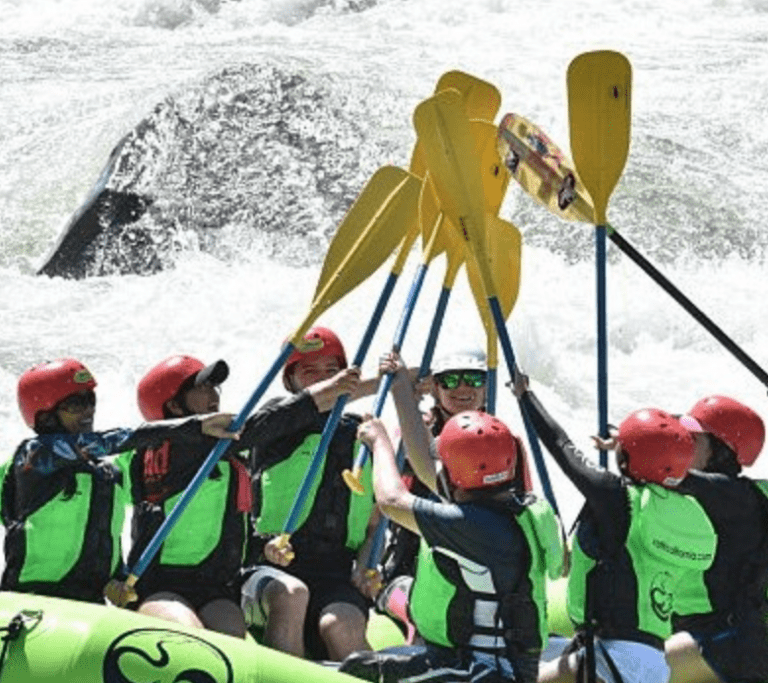 A group of people are in an inflatable rafting boat with whitewater rapids behind them. They are all wearing helmets and lifejackets and are holding their paddles high in the air in the center of the boat, doing a "paddle high five"