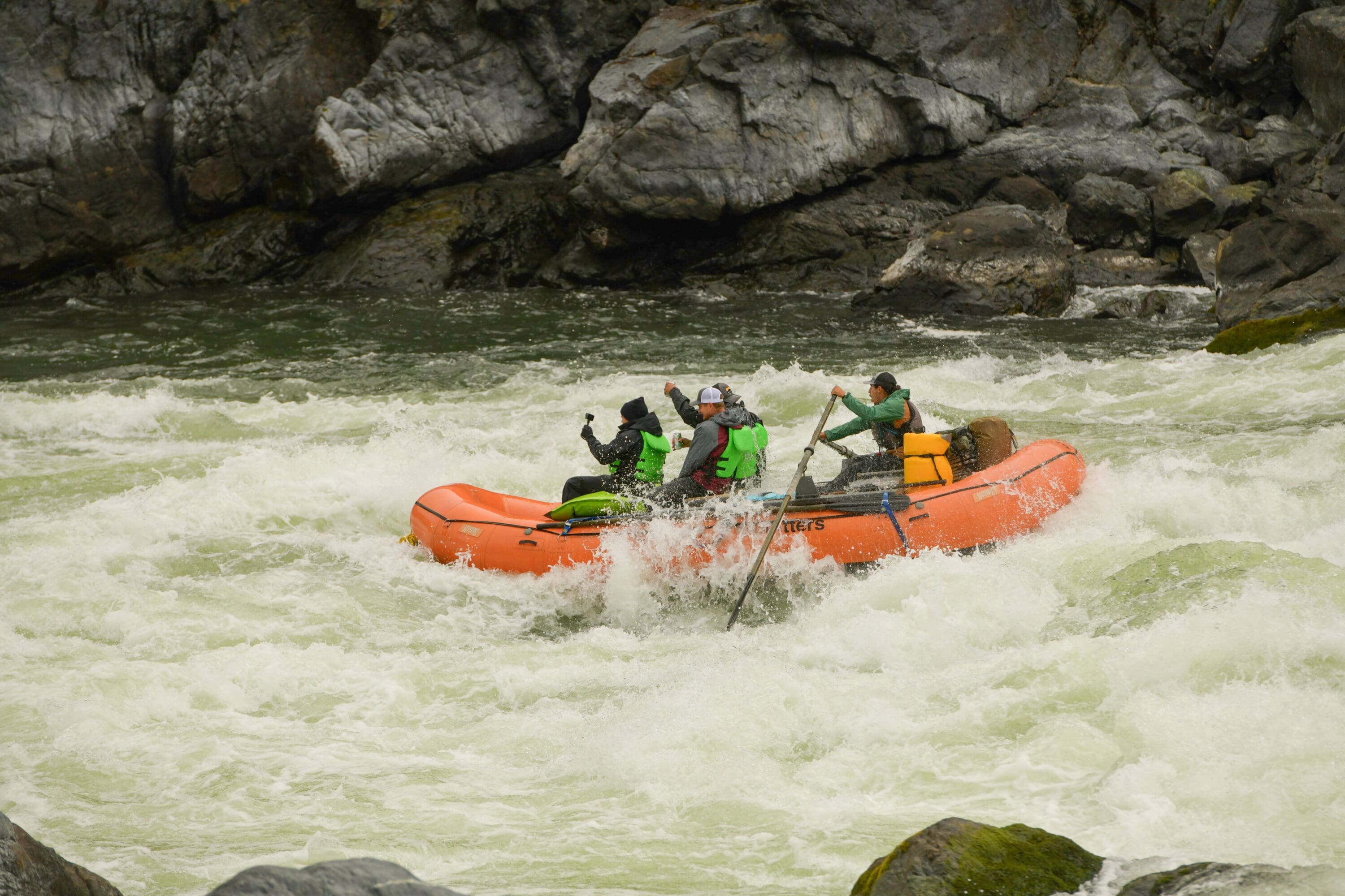 Orange inflatable boat navigating whitewater rapids on the Snake River. Everyone is wearing lifejackets and the man in the back is holding the paddles while those in the front are holding a camera.