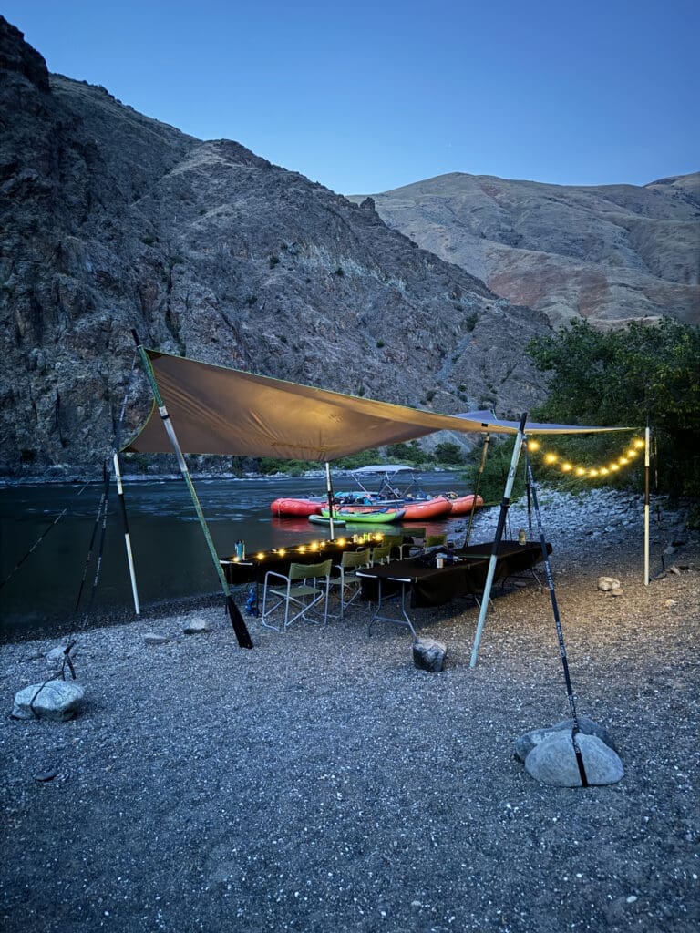 Camp set up next to calm river. A tent with tables and chairs underneath. Tables have candles lit and lights are strung up. Many inflatable boats tied up on the shore of the river.